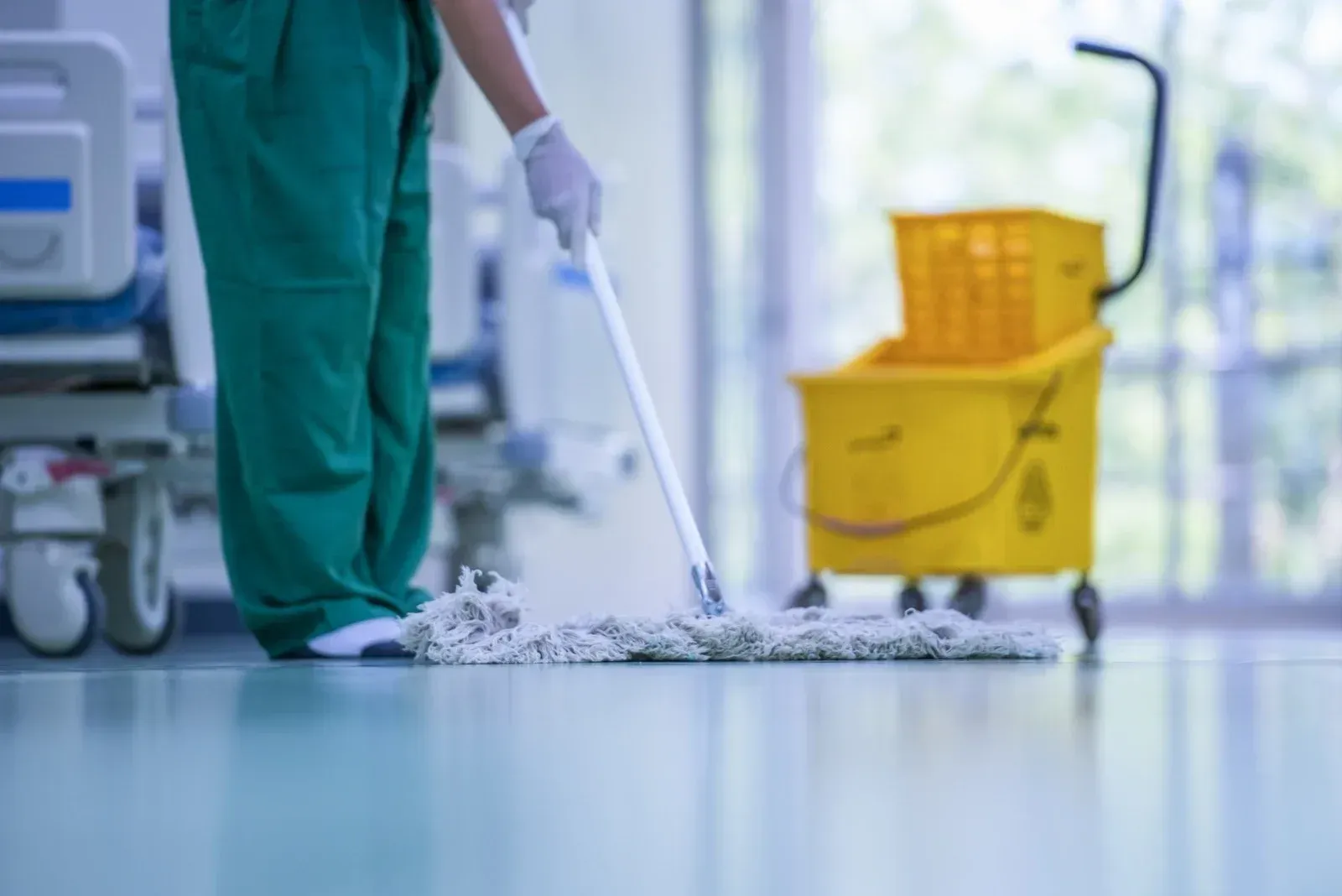 Person in green scrubs mops a hospital floor near a yellow cleaning cart and a hospital bed.