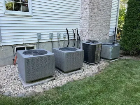 Four outdoor HVAC units beside a house, set on gravel near a stone wall and shrubs.