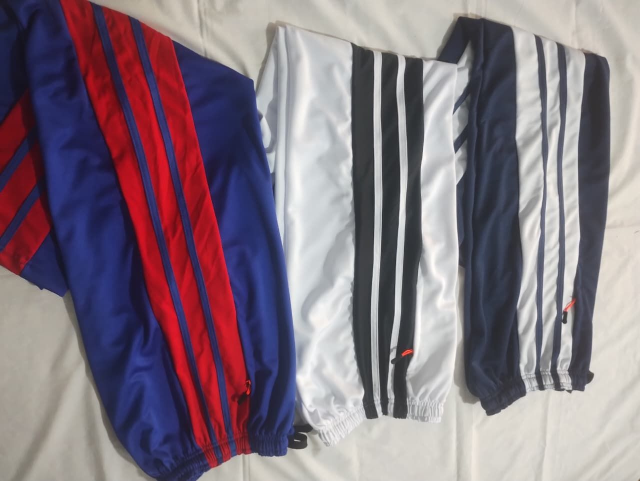 Tres pantalones deportivos Adidas: azul con rayas rojas, blanco con rayas negras y azul marino con rayas blancas.