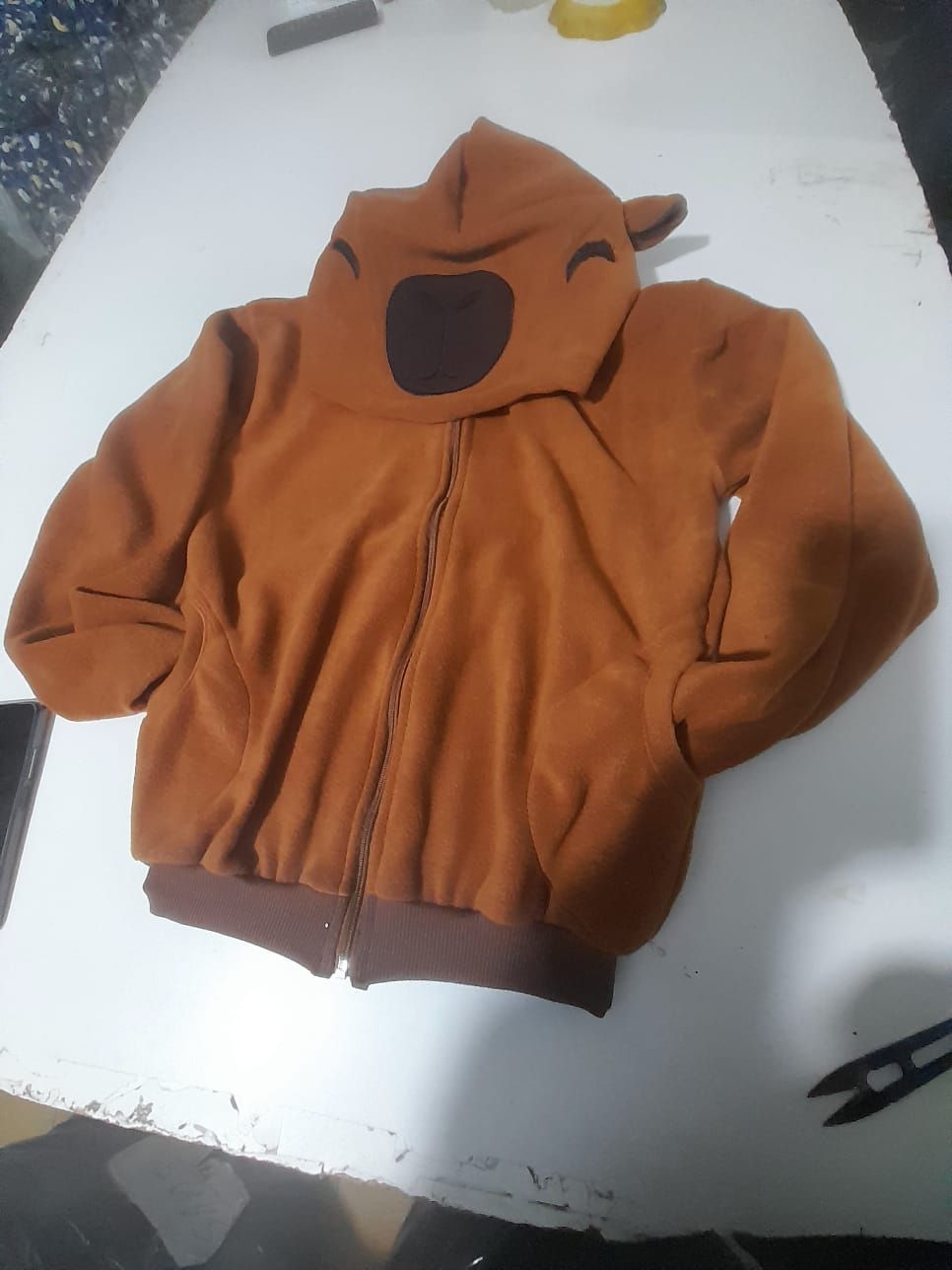 Sudadera marrón con capucha y cara de oso estampada, cerrada con cremallera sobre una superficie blanca.