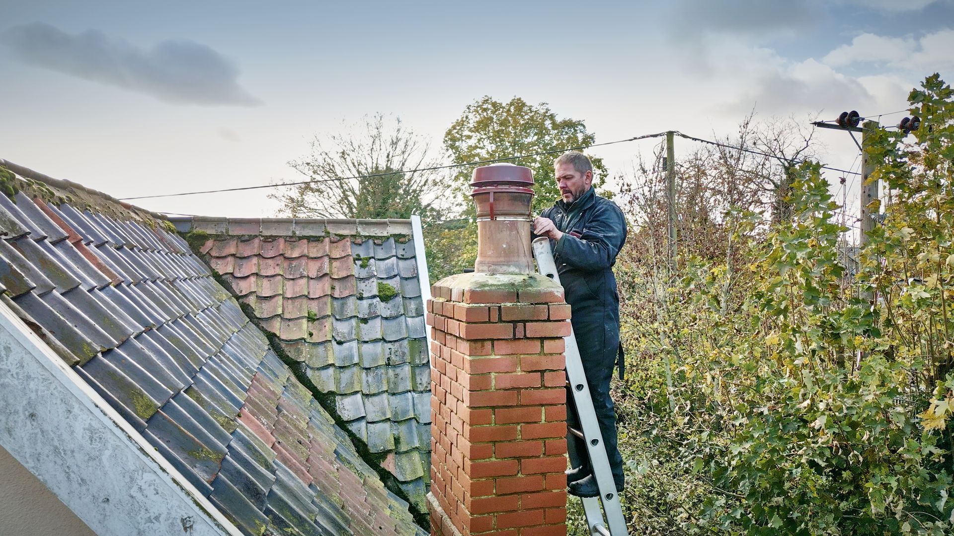 Person on a ladder inspecting a chimney on a roof with trees in the background.