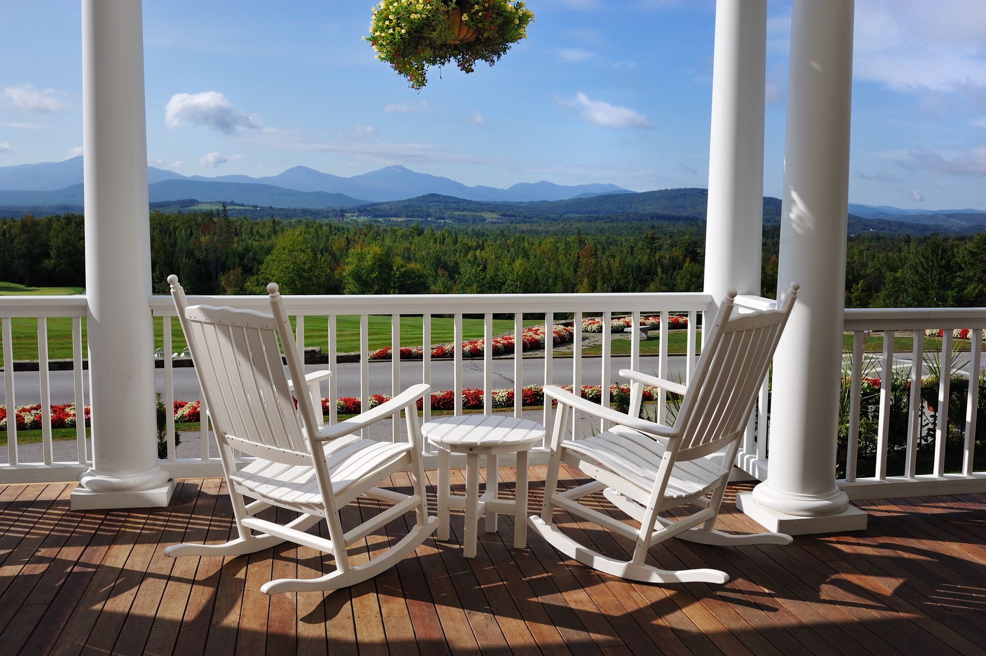 Two white rocking chairs on a porch with a small table, overlooking mountains and trees.