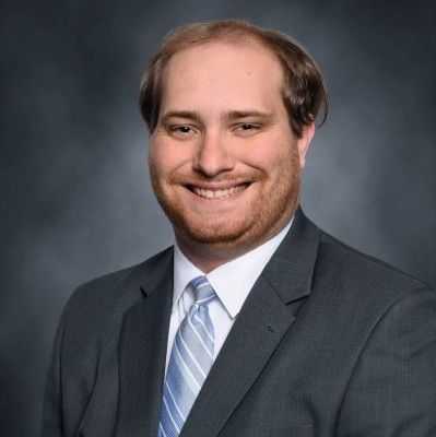 Man with gray hair and beard, wearing a suit and tie, against a blue background.