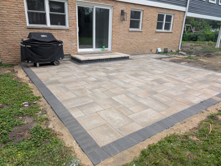 A newly constructed patio with tan Unilock pavers, dark border, and a black grill next to a brick house.