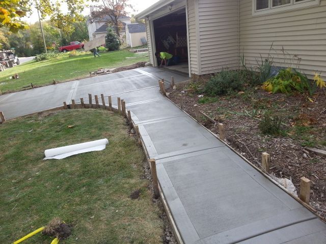 Concrete driveway and walkway being poured next to a house with a garage and grass lawn.