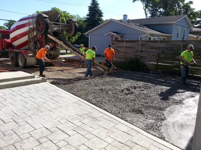 Construction workers pouring concrete from a truck into the driveway area, near a stamped concrete patio.