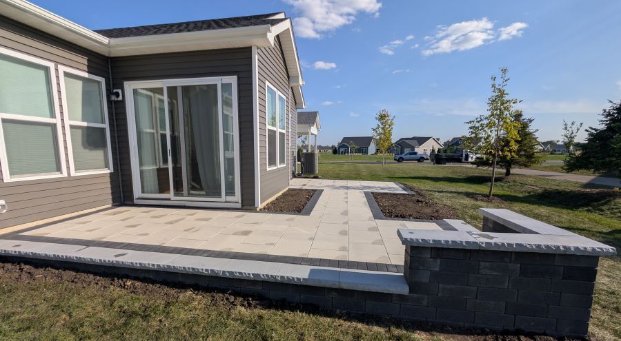 Patio with light pavers and dark gray retaining wall, next to a house with sliding glass doors.