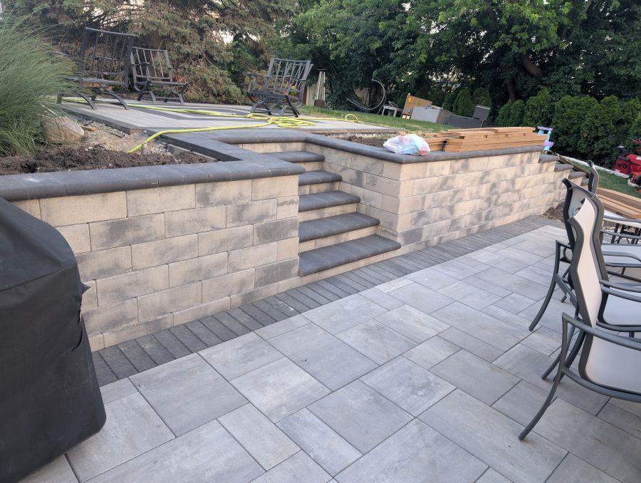 Stone patio with retaining wall, stairs, and grill. Grey pavers, neutral brickwork, outdoor setting.