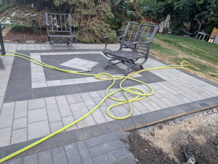 Patio with gray and black brick pavers, metal chairs, and a coiled yellow garden hose.