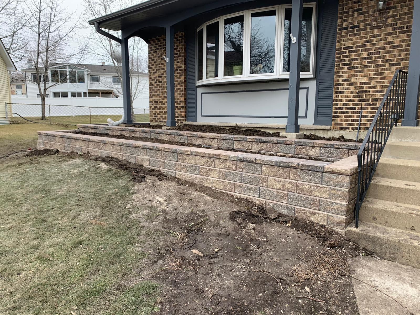 A brick retaining wall in front of a house, with a porch and steps leading up. Brown and gray brick.