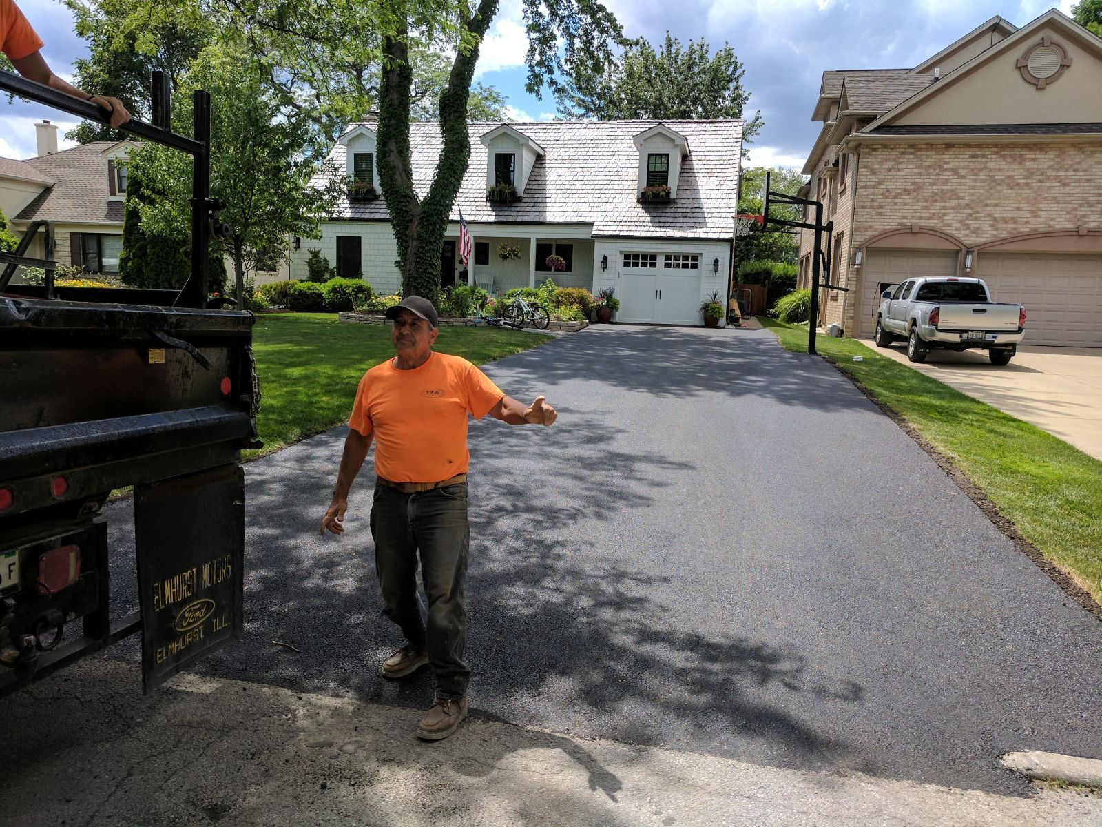 Man in orange shirt gestures at freshly paved driveway. Truck and homes in background.