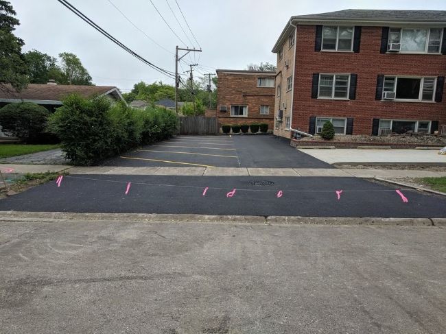 Newly paved parking lot in front of a brick apartment building; pink flags mark a line across the asphalt.