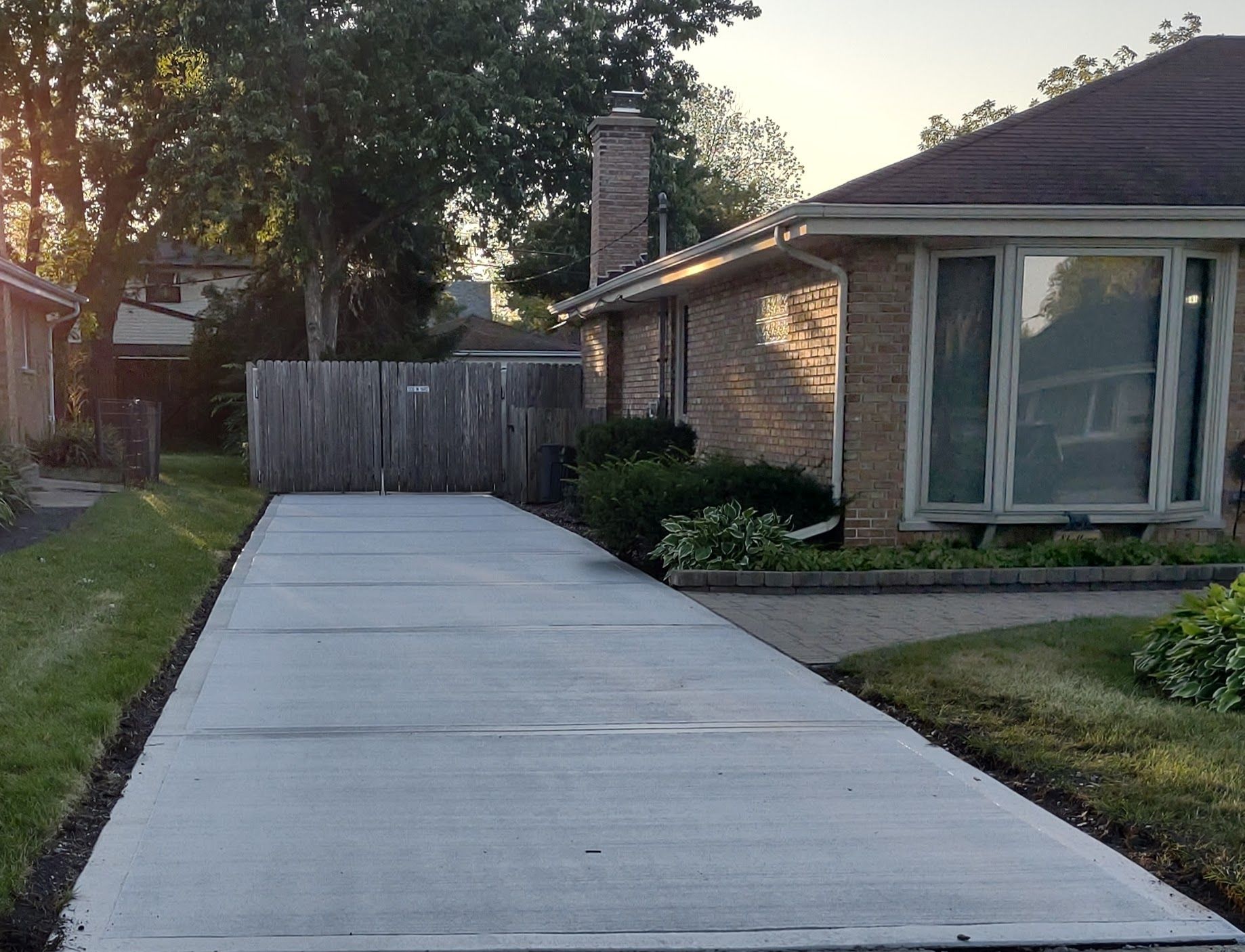 Newly paved concrete driveway leading to a brick house with large windows and a wooden fence.