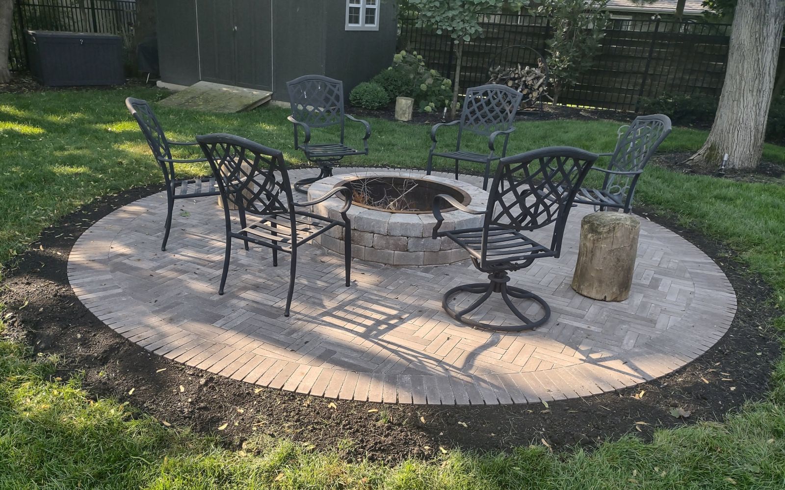 Round brick patio with a fire pit, surrounded by black metal chairs on green grass.