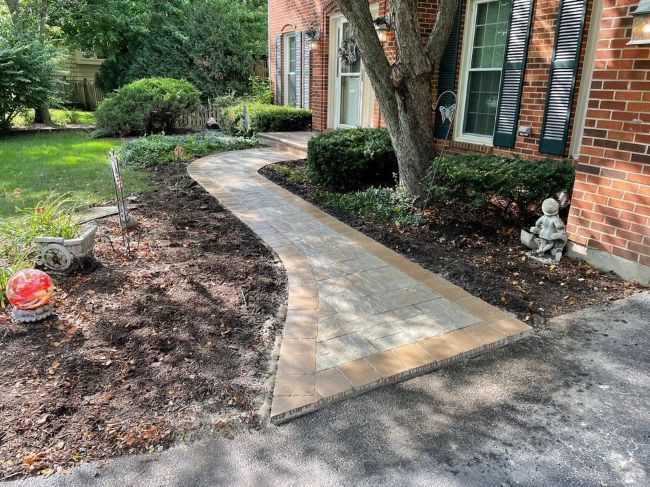 A brick paver walkway curves towards a brick house. Mulch and plants border the path.