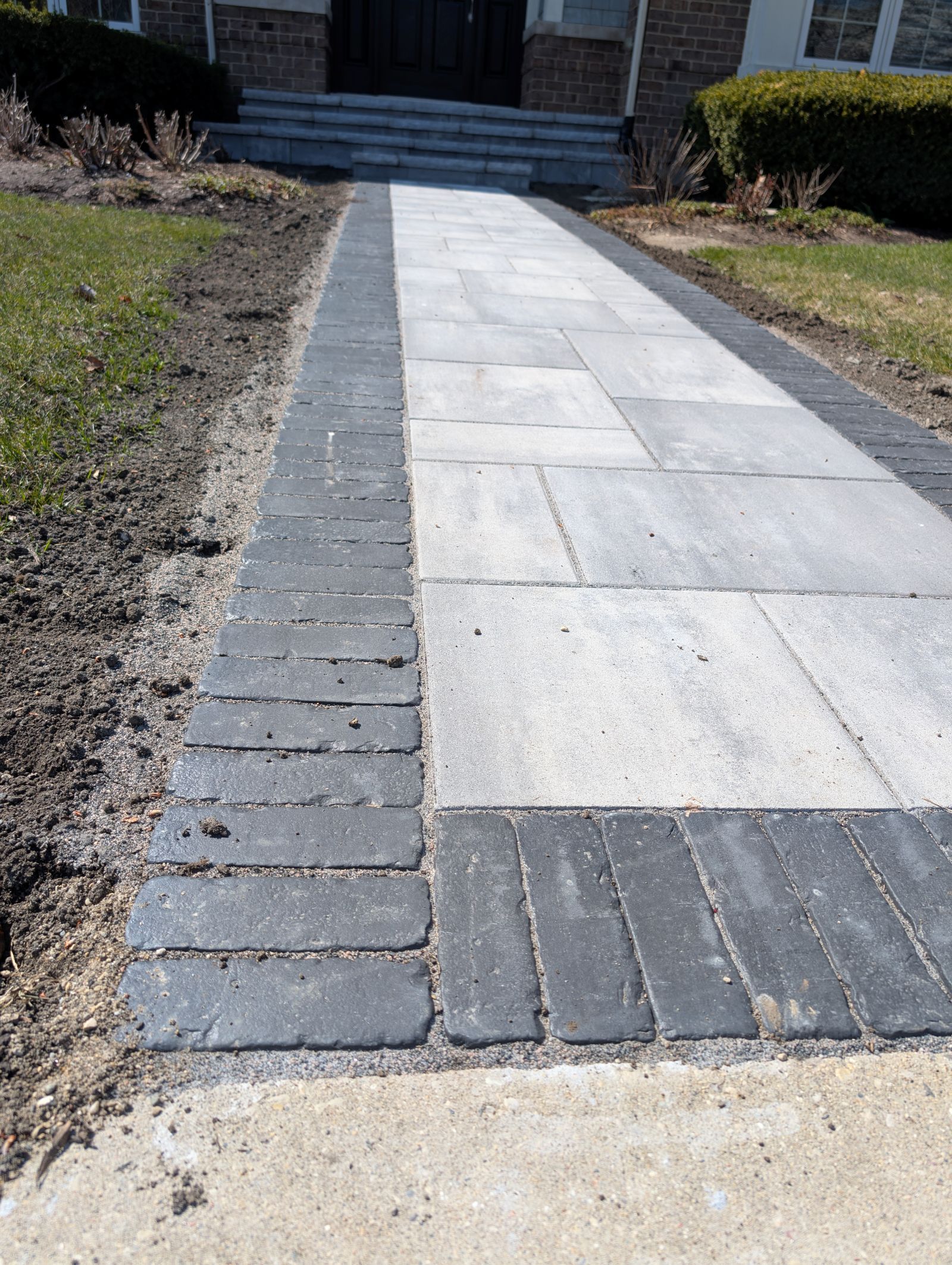 Paved walkway of grey rectangular pavers with dark grey brick border, leading to house steps.