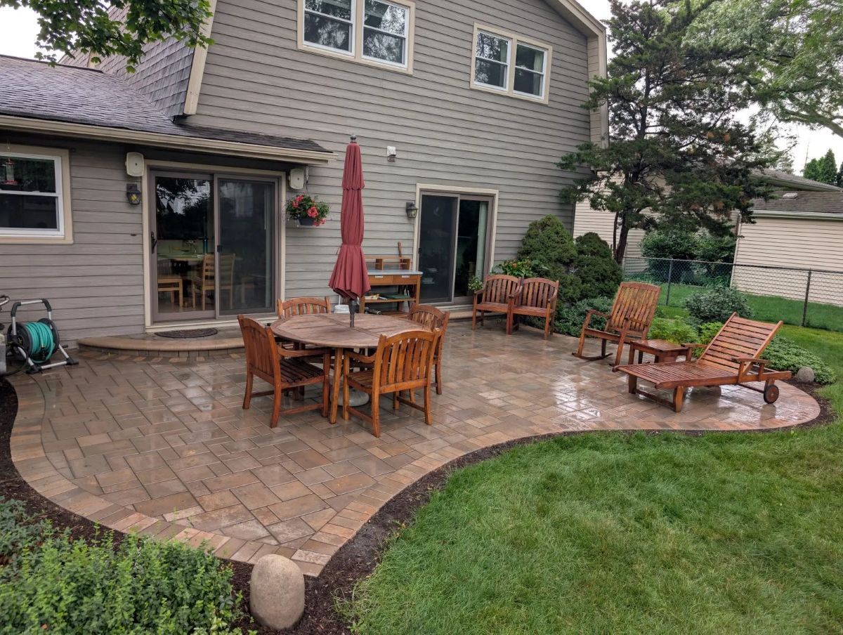 Patio with wooden furniture, umbrella, and lush green lawn next to a two-story house.
