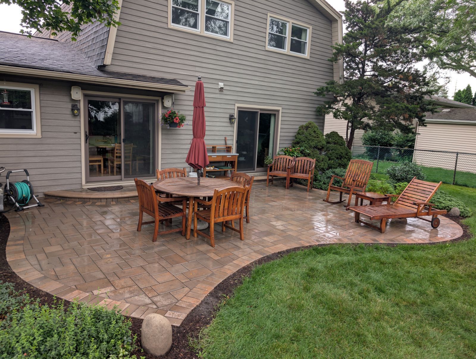 Patio with wooden furniture, umbrella, and lush green lawn next to a two-story house.