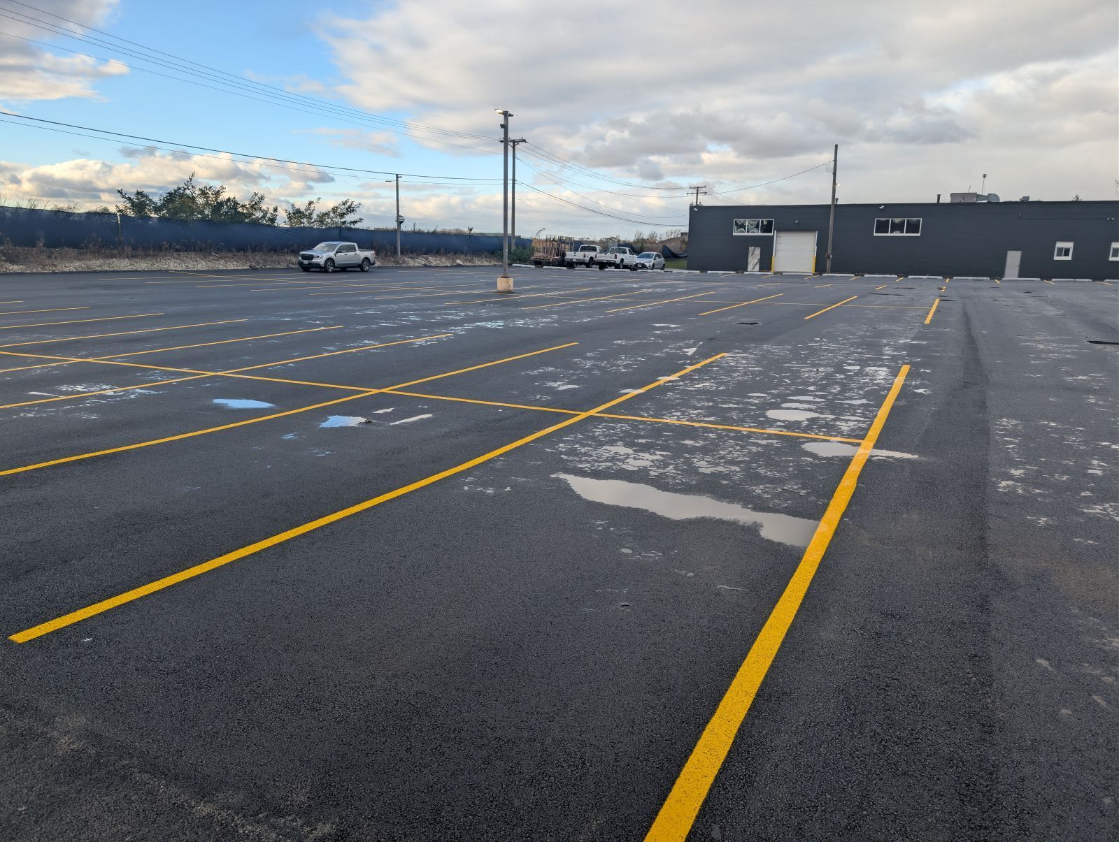 Newly paved parking lot with yellow lines, a few vehicles, and a gray building under a cloudy sky.