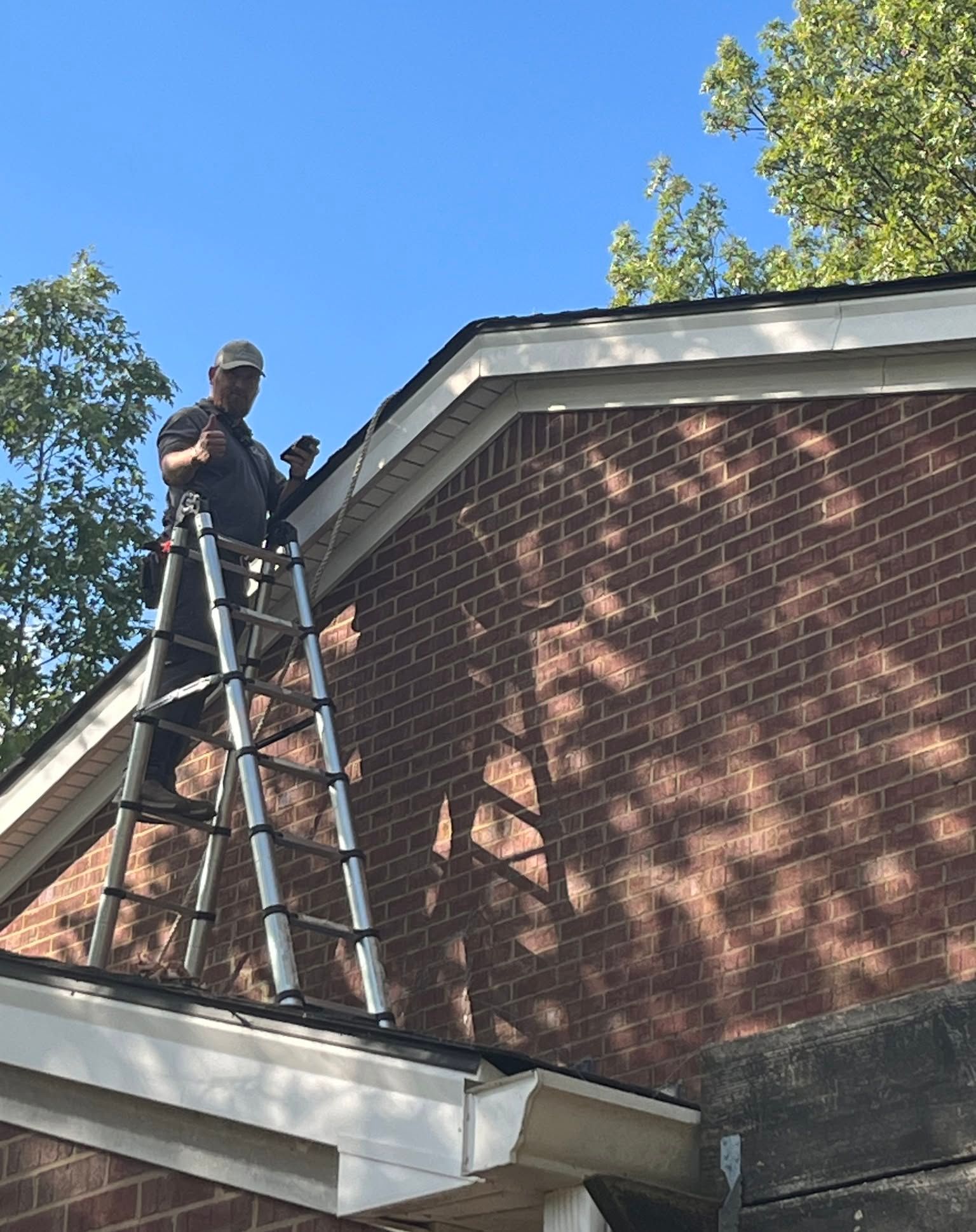 Man on a ladder repairs roof's edge on a brick building under a blue sky.
