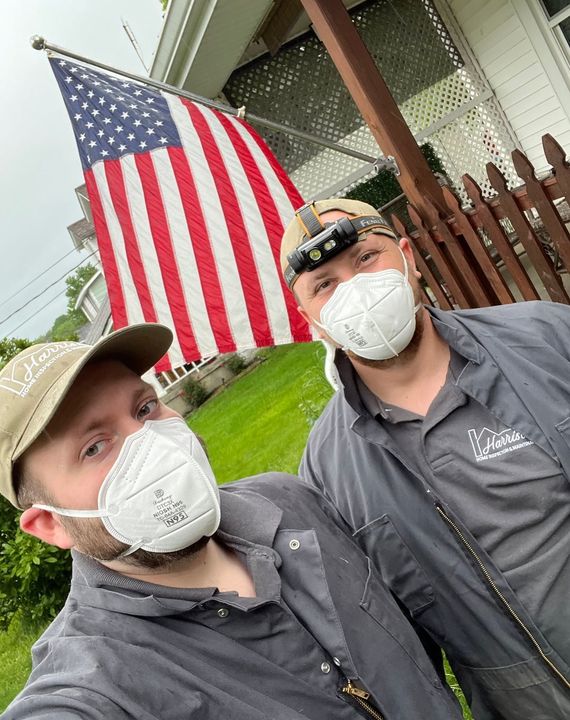 Two men in work clothes and masks pose with an American flag in the background.