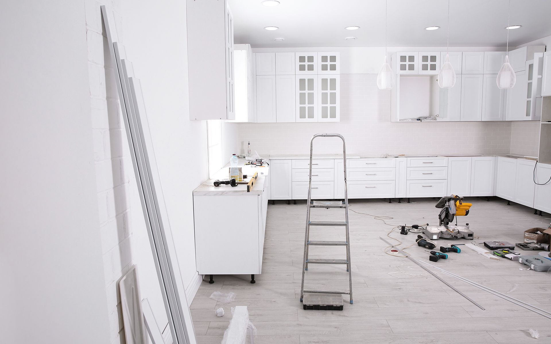 White kitchen under renovation with cabinets, ladder, and tools scattered on the floor.