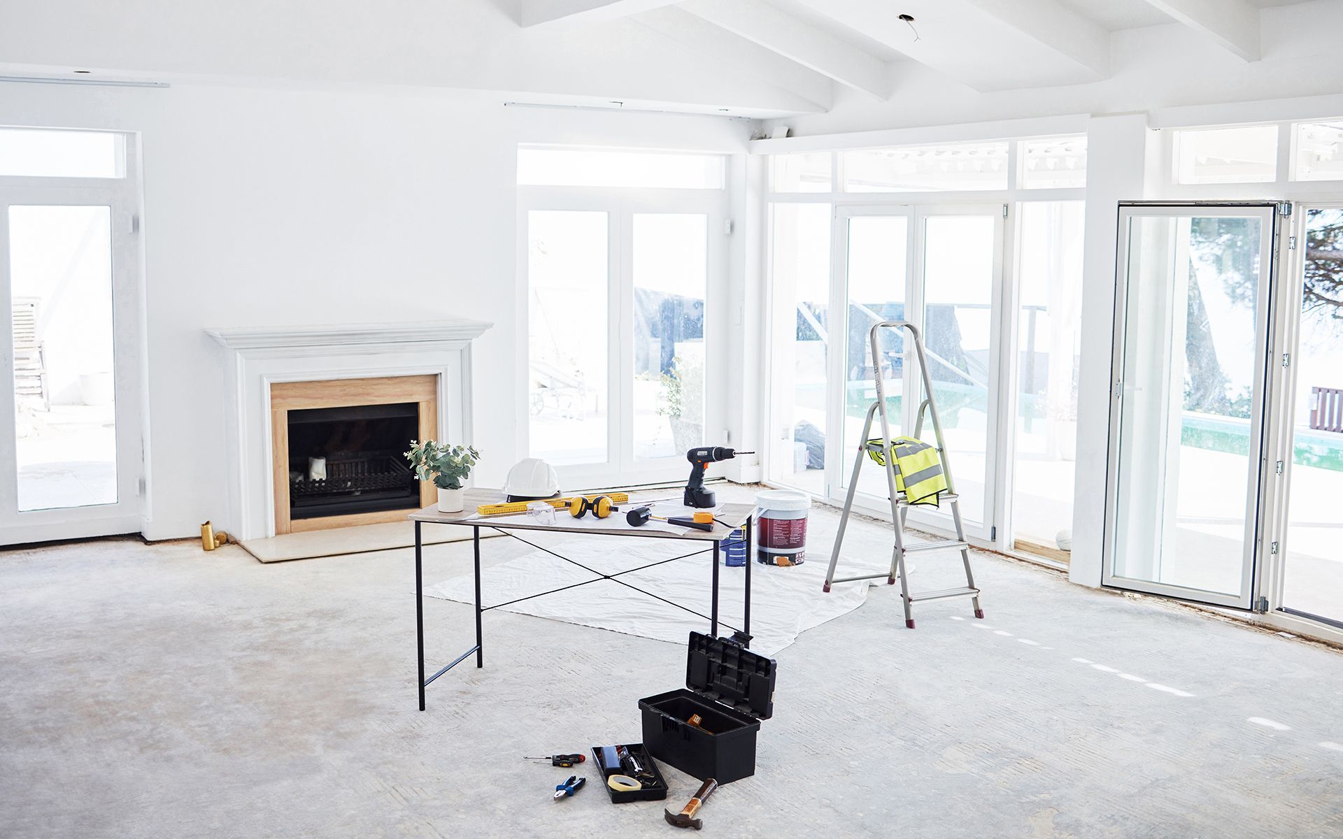 White-walled room under renovation with tools on a table, ladder, fireplace, and large windows.
