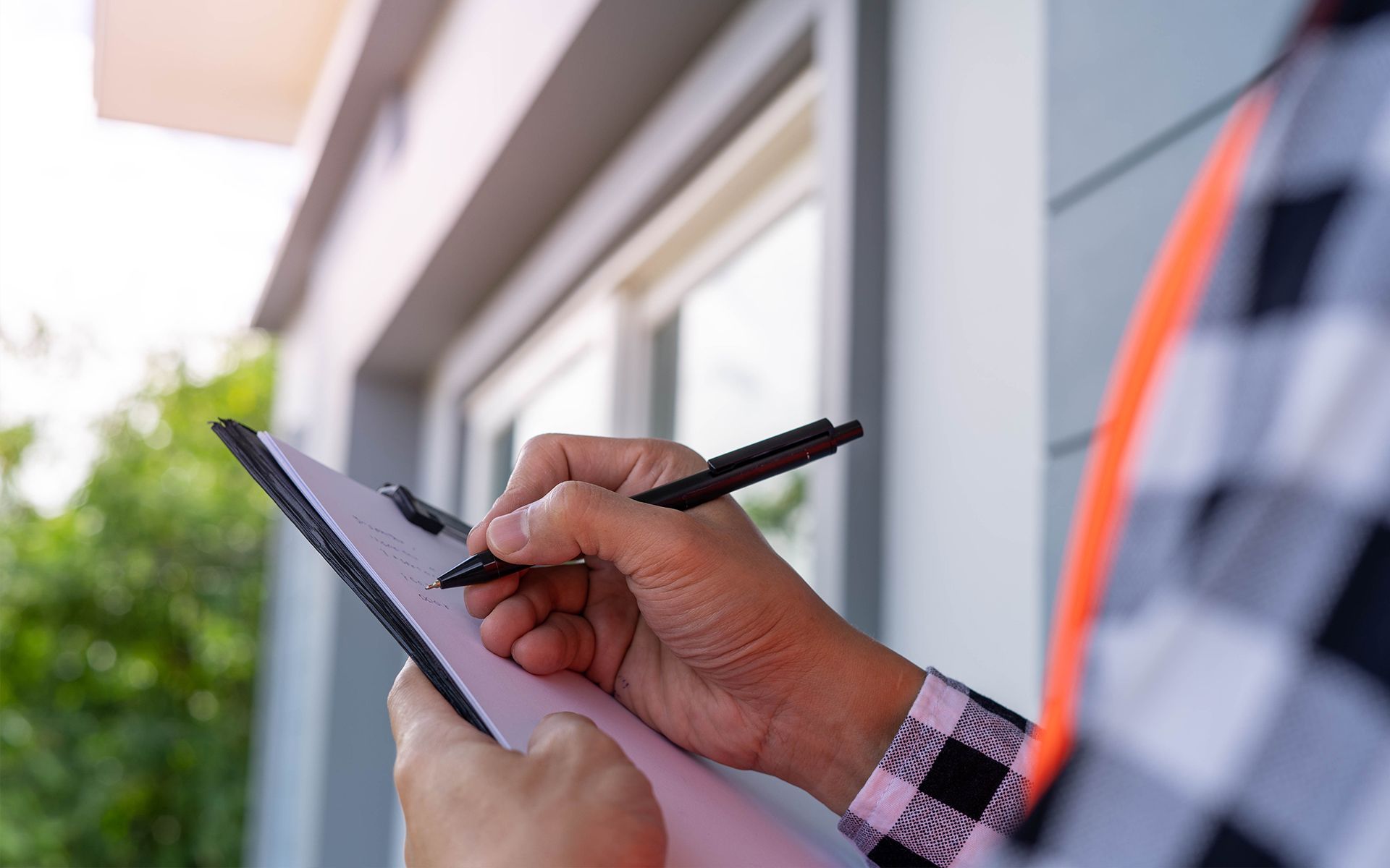 Person in plaid shirt taking notes on a clipboard next to a house; outdoor setting.