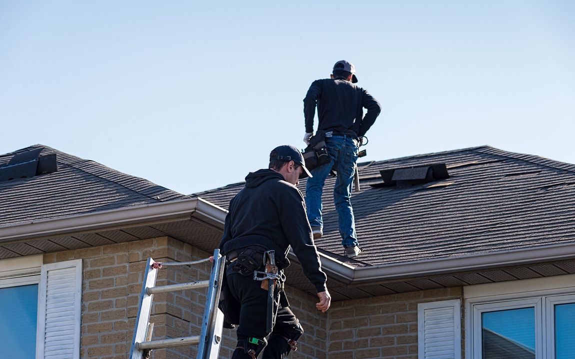 Two roofers working on a shingled roof on a sunny day; one on a ladder, one on the roof.