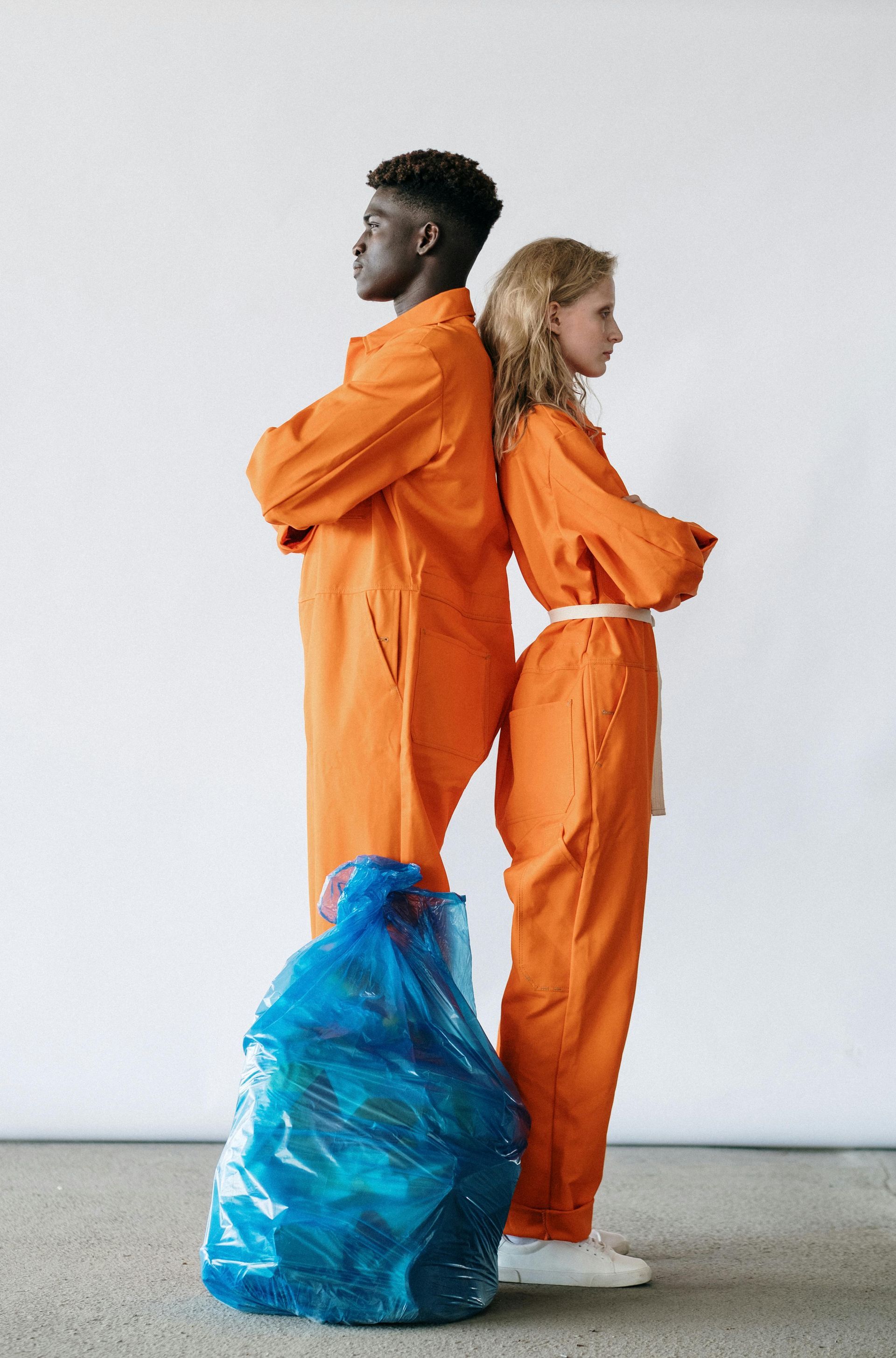 Two people in orange jumpsuits stand back-to-back, arms crossed, beside a blue trash bag against a white wall.