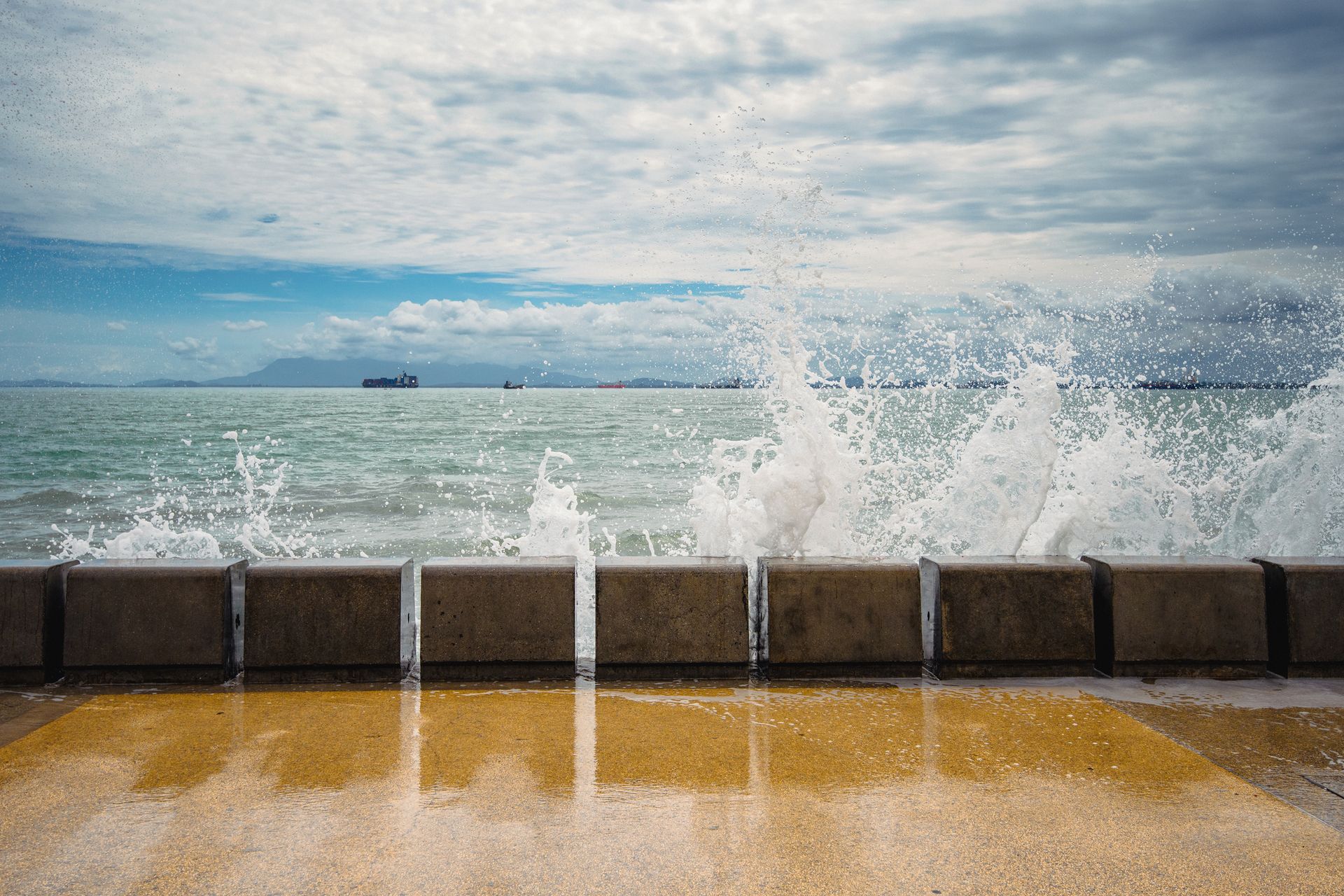 Ocean waves splashing against a seawall under a cloudy sky.