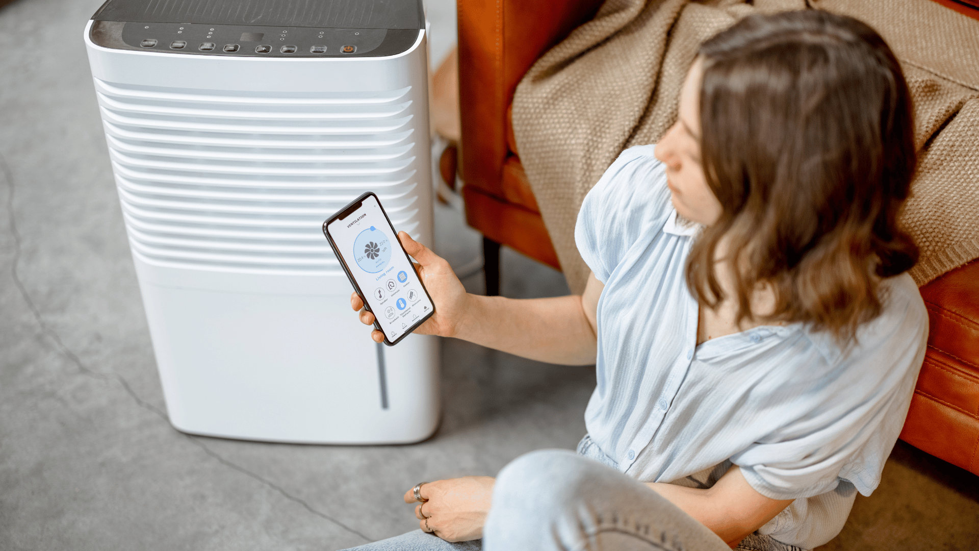 A woman is sitting on the floor holding a cell phone in front of a dehumidifier.