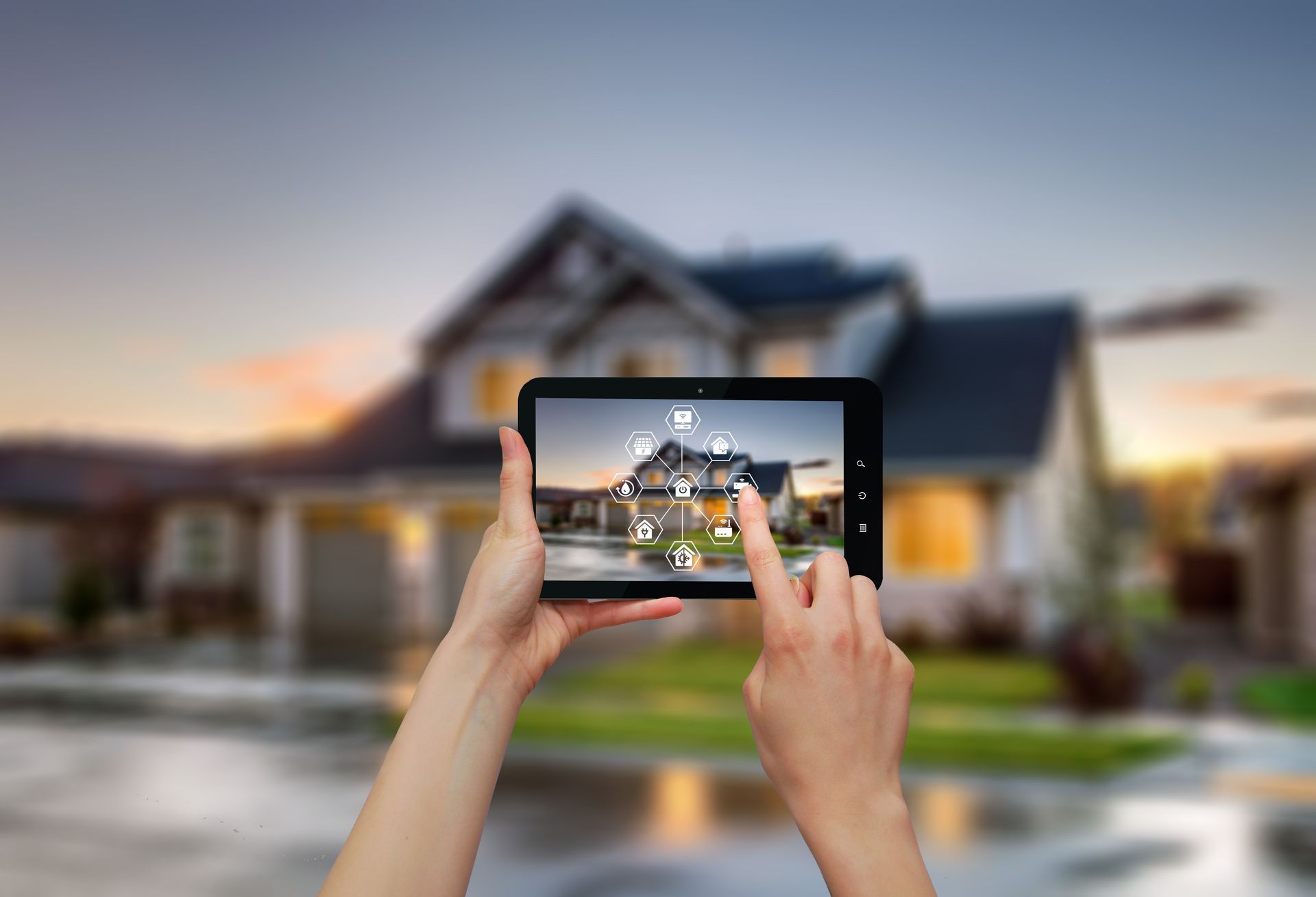Hands holding a tablet displaying a smart home interface over a blurred view of a house at twilight.