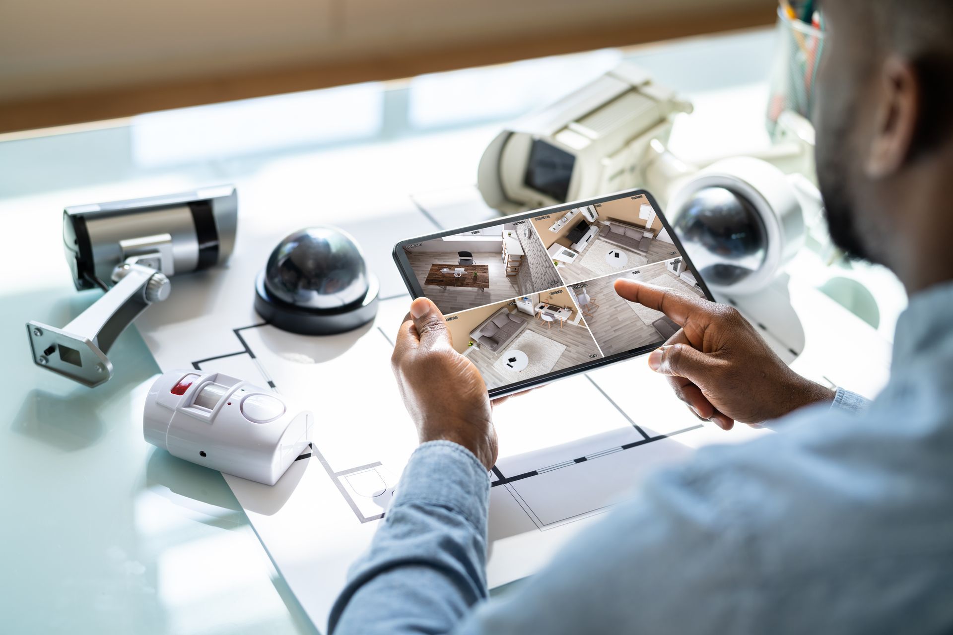 A person reviews security camera feeds on a tablet while sitting at a desk with various surveillance hardware and blueprints.
