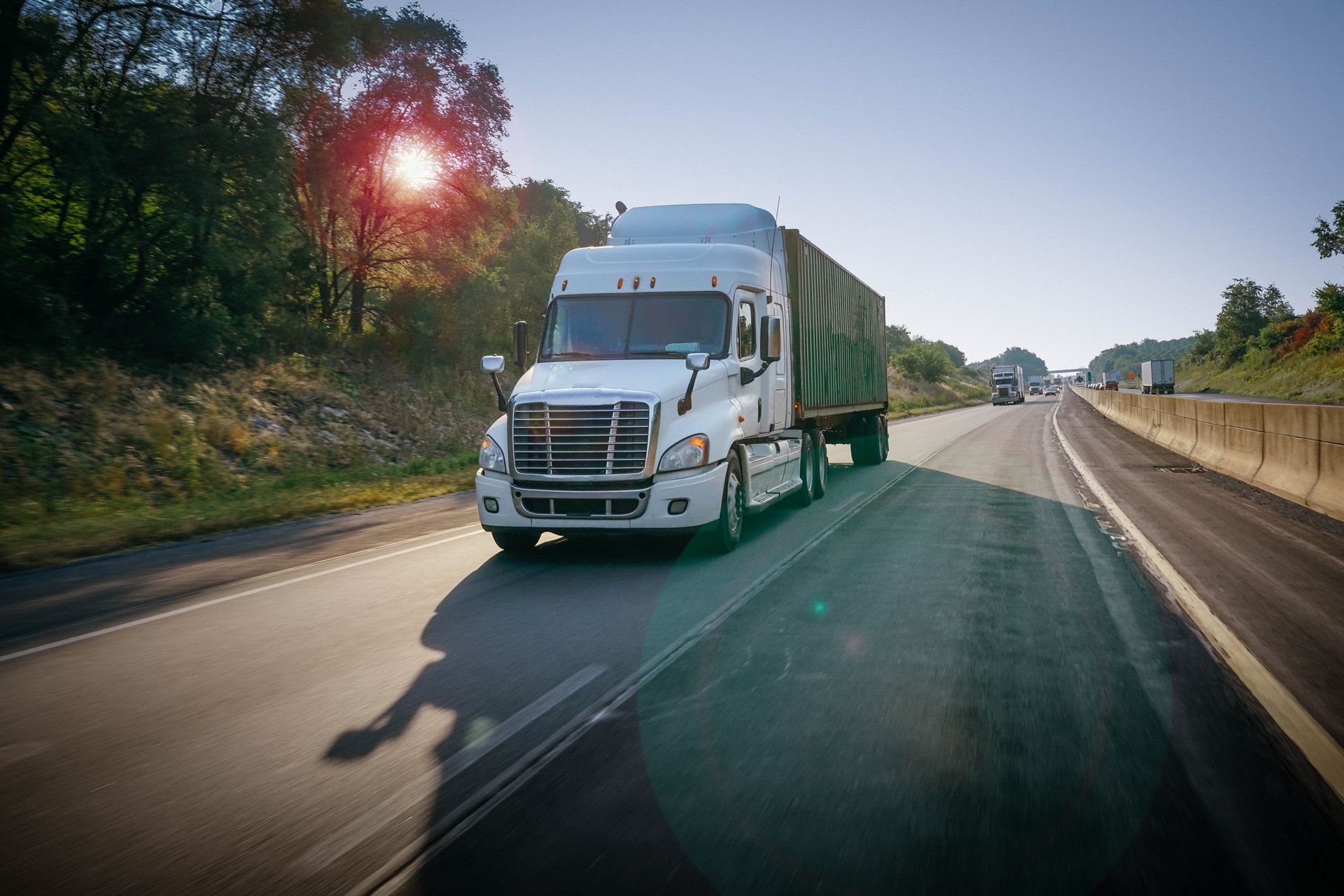 White semi-truck driving on a highway, green trees and blue sky visible.