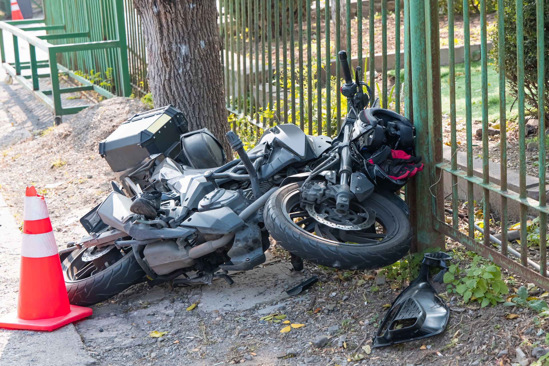 Motorcycle crashed into a fence next to a tree, with an orange traffic cone nearby.