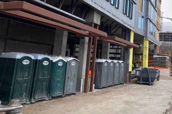 Row of portable toilets on a construction site, beneath a brown awning; building in background.