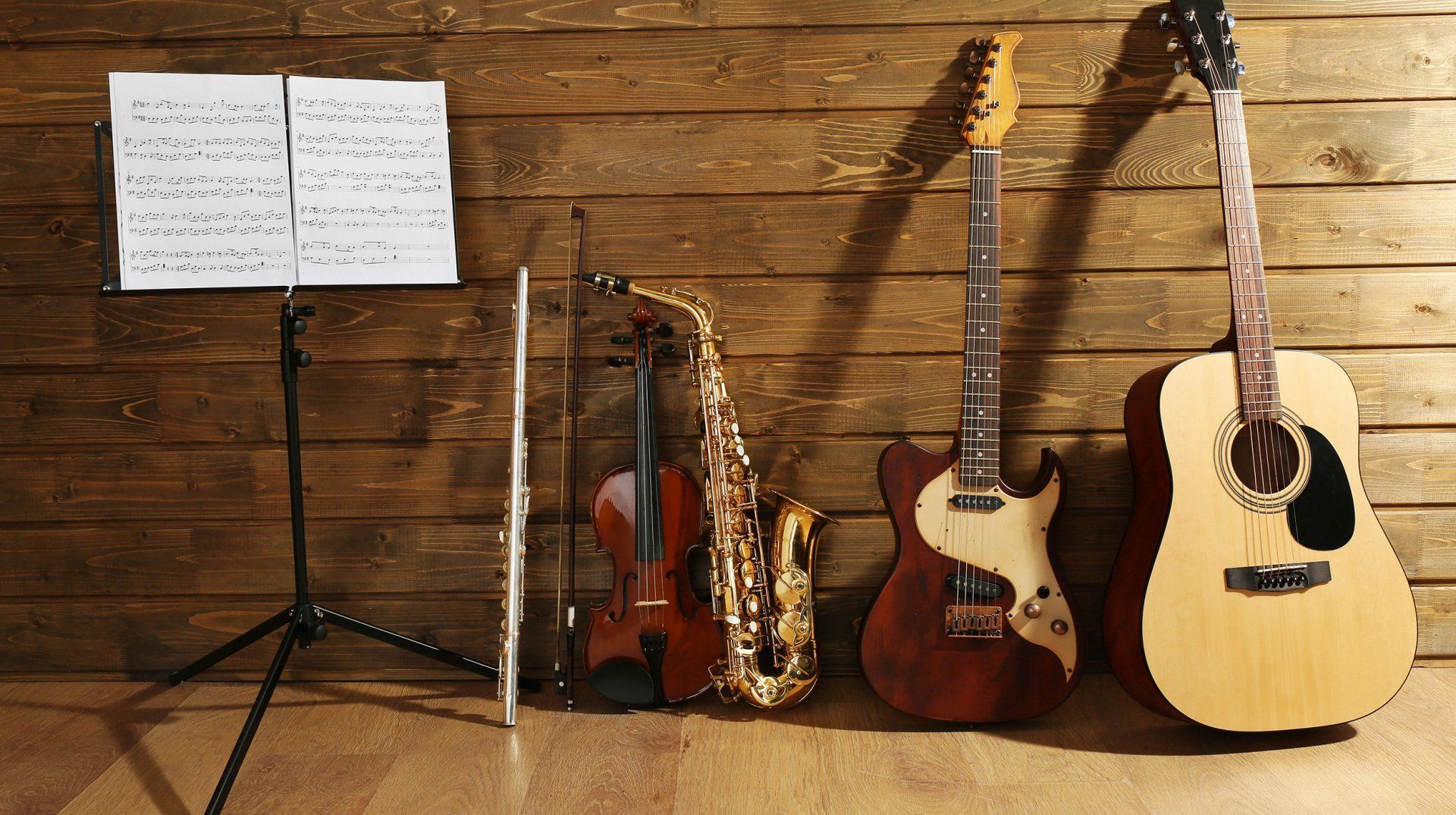 A violin, saxophone, guitar, and flute are sitting in front of a wooden wall.