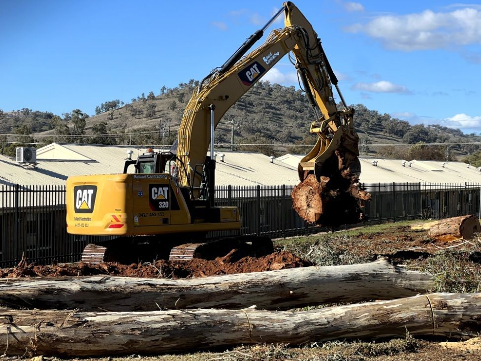 Excavator removing tree stump at a demolition site near a fenced building, with hills behind.