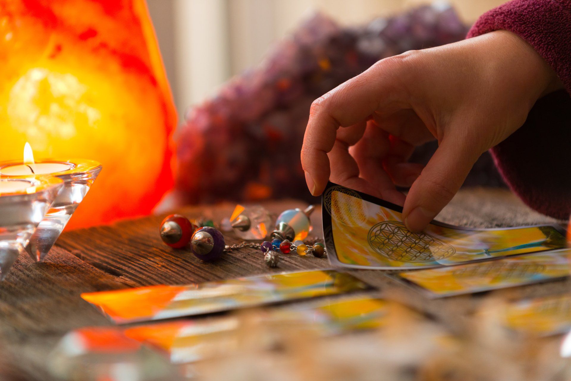 La mano de una persona seleccionando una carta del tarot sobre una mesa de madera con velas y cristales.