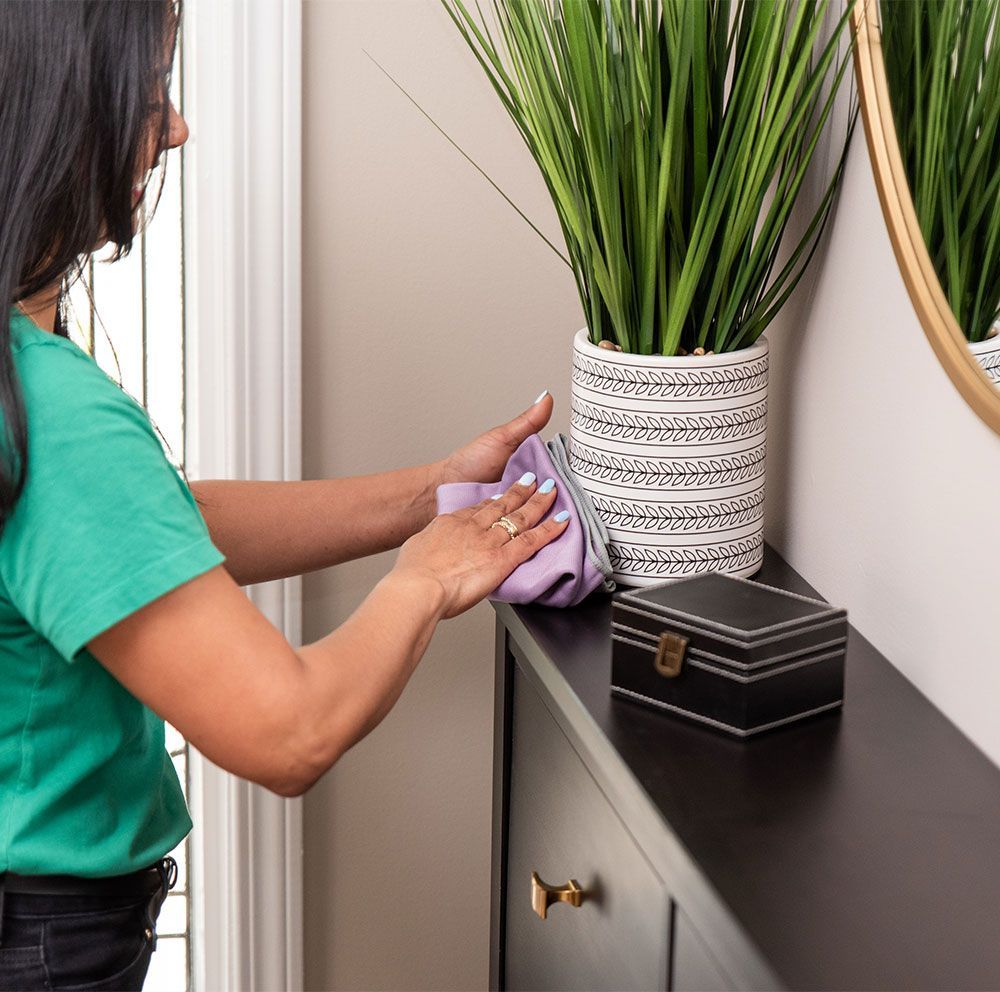 Woman wiping a dark dresser with a purple cloth; a potted plant and box are on top.