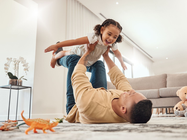 Man lying on floor, lifting child. Both are smiling, in a bright living room with toys.