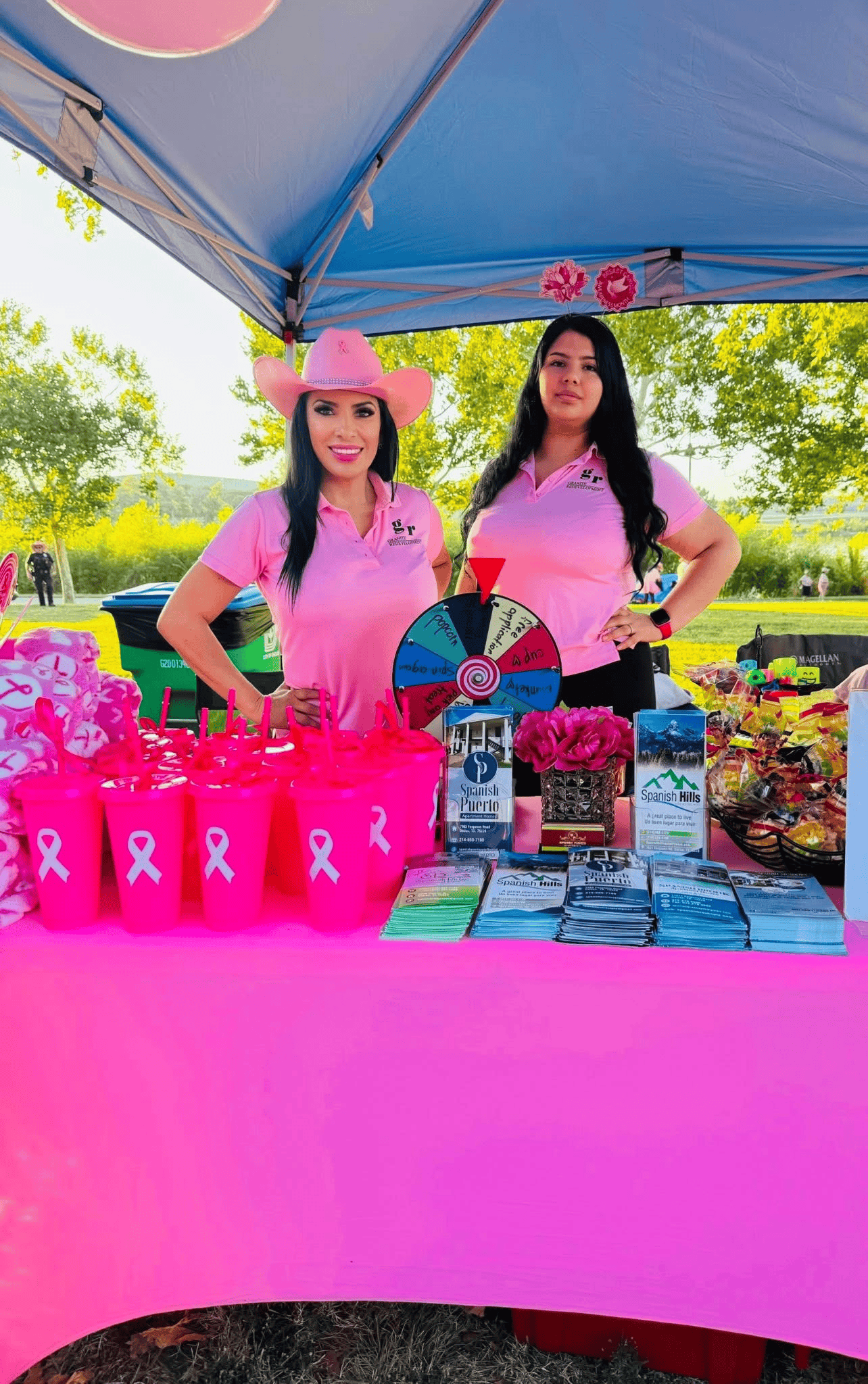Two women at a pink table promoting breast cancer awareness, wearing pink shirts and a cowboy hat.