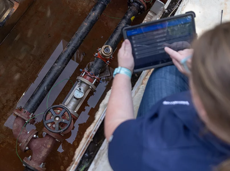 A person using a tablet to inspect pipes in a trench filled with brown water.