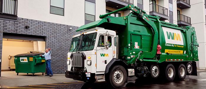 A Waste Management truck next to a building, with a person moving a dumpster.