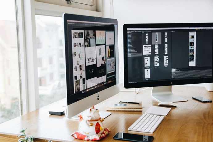 Two computer monitors are sitting on a wooden desk next to a window.