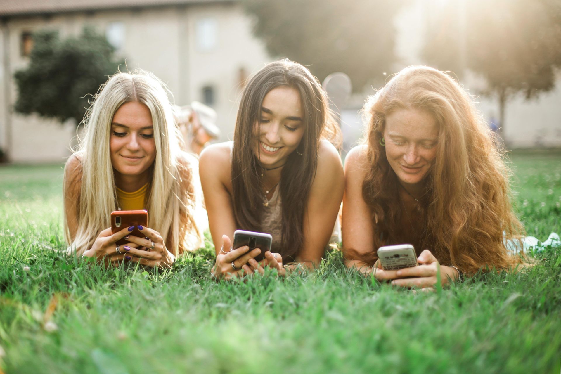 Three women are laying on the grass looking at their phones.