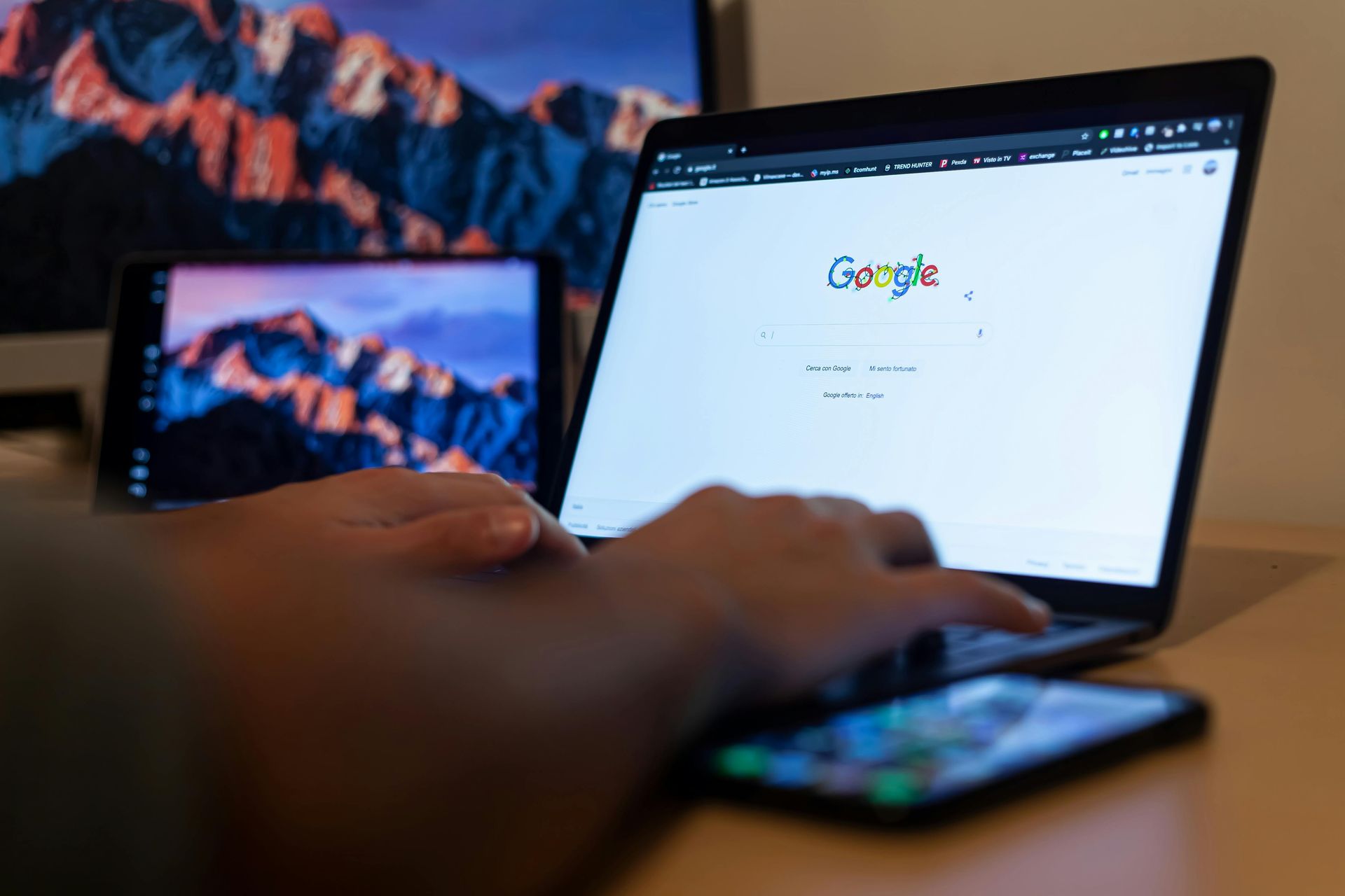 A person is typing on a laptop computer while using a cell phone.
