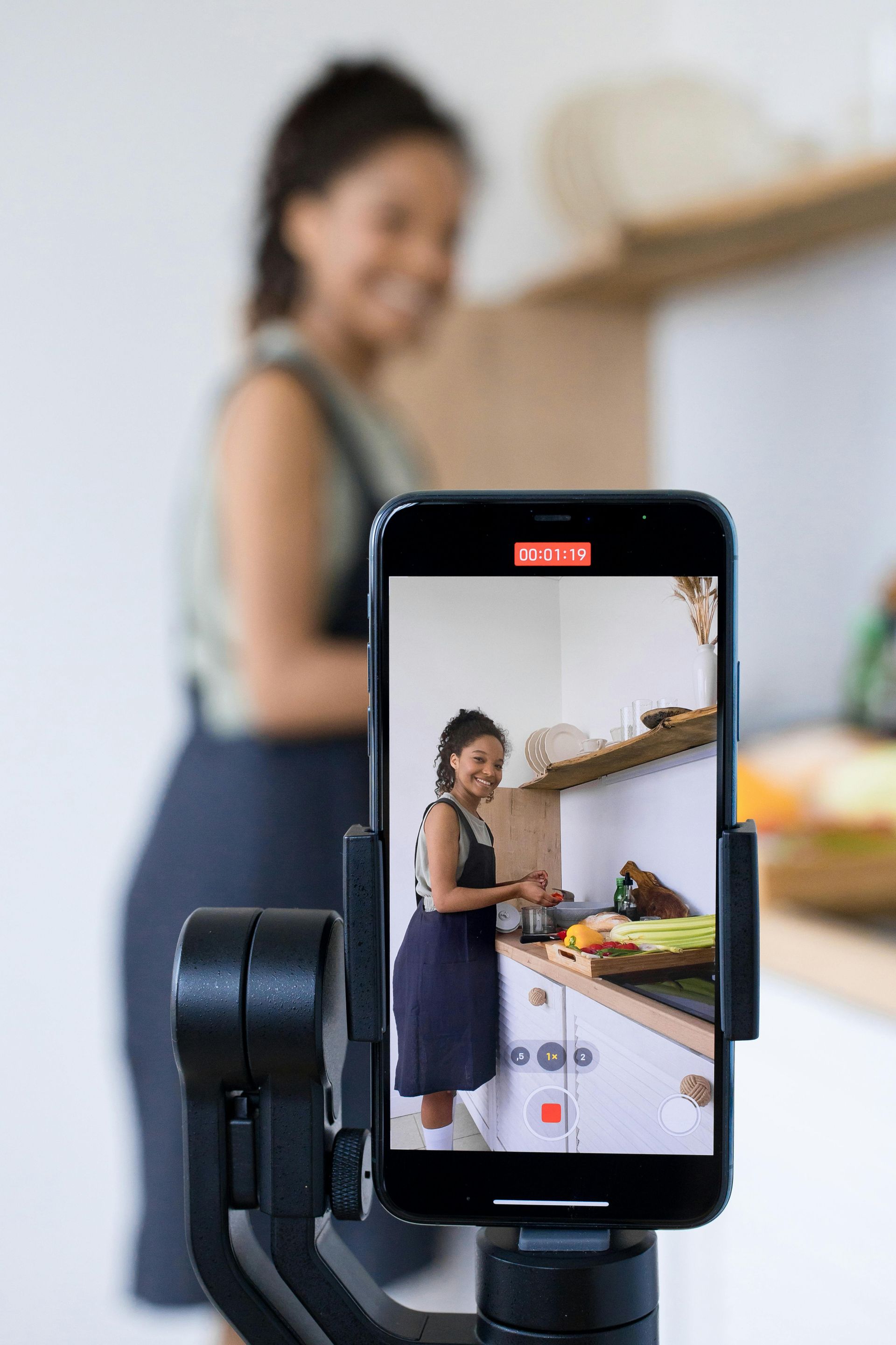 A woman is cooking in a kitchen while being filmed on a cell phone.