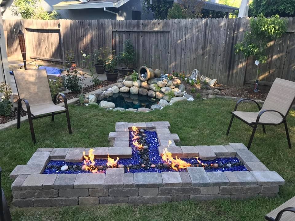 Fire pit with blue glass and flames in backyard with pond and chairs.